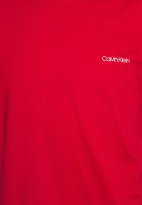 Červené bavlněné tričko s kulatým výstřihem a malým bílým logem "Calvin Klein" na horní levé straně. Hladký povrch.