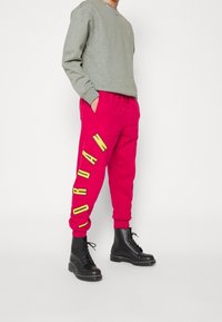 Persona vestida con pantalones de chándal rojos con el texto amarillo "JORDAN", sudadera gris y botas negras con cordones, de pie contra un fondo blanco.