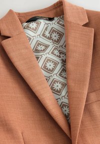 Blazer marrón con una tela texturizada; cuenta con solapas y un forro interior con patrones geométricos en azul claro y marrón.
