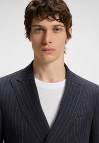 Marineblau gestreifter Blazer mit Revers und Taschenakzenten, über einem weißen strukturierten Rundhals-Shirt getragen.