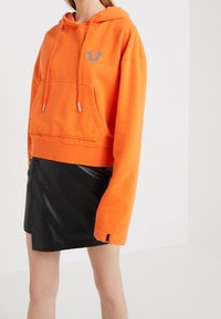 Sweatshirt à capuche orange avec une poche avant, logo gris et cordon de serrage. Assorti à une mini-jupe en simili cuir noire.