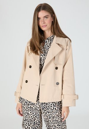 Beige double-breasted jacket with black buttons, wide lapels, and adjustable cuffs, worn over a black and beige leopard-print shirt.