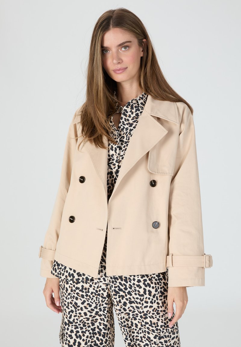 Beige double-breasted jacket with black buttons, wide lapels, and adjustable cuffs, worn over a black and beige leopard-print shirt.