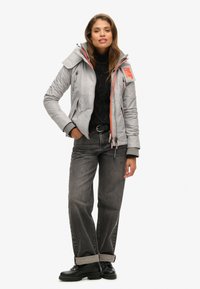 Veste isolante grise avec un col haut, des accents orange et des détails zippés, associée à un pull noir, un jean gris et des bottes noires.