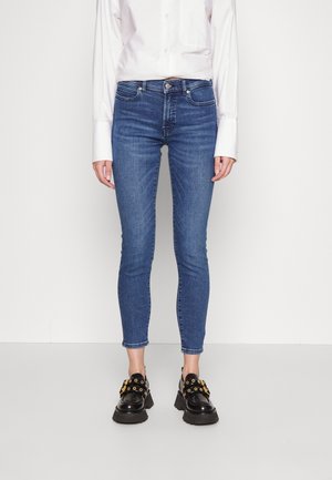 Femme portant un jean slim bleu, une chemise blanche à manches longues et des mocassins noirs épais avec œillets dorés, debout sur un fond clair uni.