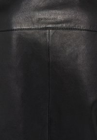 Schwarze Lederjacke mit glatter Textur, minimalistischer Gestaltung und dem geprägten Logo "TRUSSARDI" auf der Rückseite nahe dem Kragen.