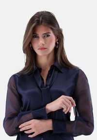 Marineblaue Bluse mit transparenten langen Ärmeln, Knopfleiste vorne, Kragen und optionalem Bindeband am Hals. Glattes Material mit glänzender Oberfläche.