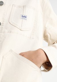 Närbild av en hand i framfickan på en krämfärgad Lee-arbetsjacka med synliga knappar och logotyp.