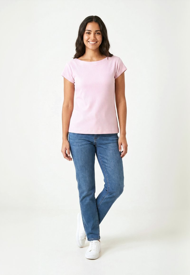 Femme debout, souriante, portant un haut à manches courtes rose clair, un jean bleu et des baskets blanches sur un fond clair uni.