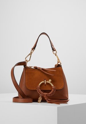 Sac à main en cuir marron avec rabat en daim, anneau doré et détail pompon, courte poignée supérieure et longue bandoulière réglable sur surface d’exposition blanche.