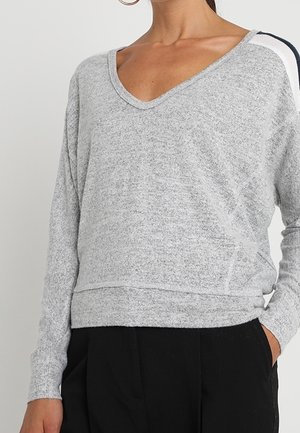 Femme portant un pull gris clair à manches longues avec col en V, pantalon noir, main dans la poche, et grands anneaux d'oreilles visibles.