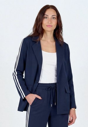 Femme aux longs cheveux bruns portant un blazer bleu marine et un pantalon assorti à rayures blanches, associé à un haut blanc, debout devant un fond blanc.