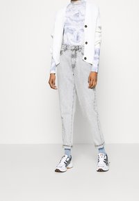 Cárdigan de punto blanco sobre una camisa de manga larga de patrón azul y blanco, combinado con jeans de talle alto gris claro y zapatillas deportivas blancas.