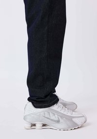 Zapatillas deportivas blancas con un diseño texturizado y detalles plateados, combinadas con pantalones de mezclilla oscuros de pierna recta y dobladillo vuelto.