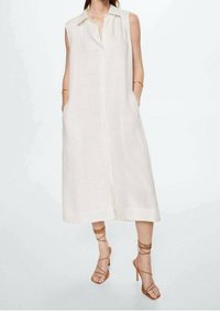 Robe sans manches, à boutons, en lin léger crème, avec des poches latérales et une silhouette droite. Accompagnée de talons beiges à lanières.