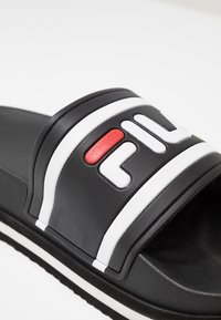 Chancla negra con una tira de cuero texturizado, que presenta un logo de FILA en blanco y rojo, así como rayas blancas en la tira y los bordes de la suela.