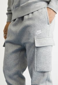 Šedé bavlněné tepláky s cargo kapsami, elastickým pasem a bílým logem Nike na levém stehně. Měkká textura, ležérní design.