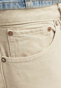 Béžové džíny z denimu s přední kapsou, měděnými nýty a viditelnou strukturou. Nosené pod modrou džínovou bundou.