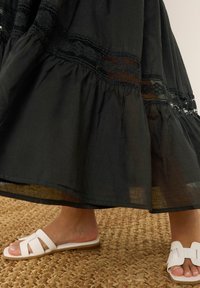 Falda negra con volantes y detalles de encaje, con una textura ligera. Combinada con sandalias blancas de punta abierta con un diseño de tiras.