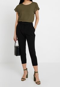 Olivgrüne Bluse mit vieleckigen Ärmeln, kombiniert mit schwarzen, knöchellangen Hosen, grünen Absatzsandalen und einer kleinen schwarzen Handtasche.