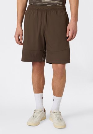 Uomo che indossa pantaloncini marrone scuro fino al ginocchio, calzini bianchi a coste e sneakers basse avorio, in piedi su sfondo bianco.
