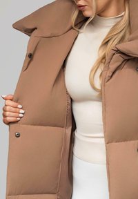 Žena s dlouhými blond vlasy, oblečená v béžových rolákových šatech, která drží rozepnutý hnědý polstrovaný kabát se cvoky.