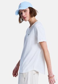 Witte T-shirt met korte mouwen en een ronde halslijn, voorzien van een geborduurd groen logo. Gecombineerd met een lichtblauwe pet en lichtgekleurde broek.