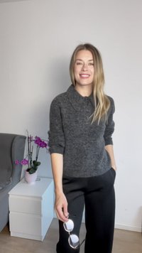 Szürke gyapjúpulóver gallérral, fekete nadrággal párosítva. Napszemüveget tart a kezében, egy fehér fiókos szekrény és orchideák mellett áll.