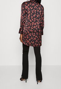 Tunique en satin noir avec imprimé léopard rouge, manches longues et coupe décontractée, associée à un pantalon noir évasé et des talons ouverts.