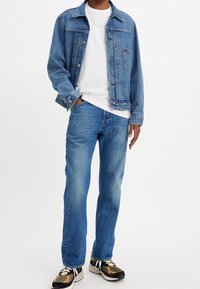 Giacca di jeans di un blu chiaro, con chiusura a bottoni, abbinata a una maglietta bianca e jeans blu coordinati. Le scarpe presentano un design decorato.