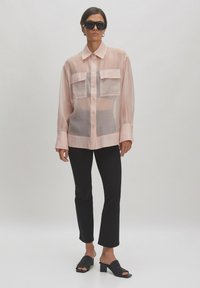 Transluzente pinkfarbene Bluse mit zwei Brusttaschen, kombiniert mit schwarzen Slim-Pants und offenen schwarzen Mules mit Blockabsatz.
