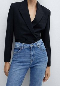 Blazer negro cruzado con solapas de pico, combinado con vaqueros de cintura alta en tono azul claro, con una textura clásica de denim.