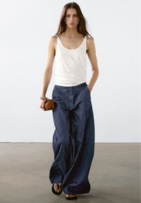 White, lightweight tank top paired with loose, dark blue denim trousers. Woven brown bag held in hand, accented with flat black sandals.