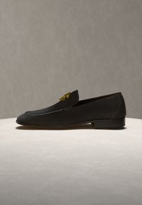 Μαύρο δερμάτινο μοκασίνι με χαμηλό τακούνι και χρυσή διακοσμητική λεπτομέρεια στο πάνω μέρος, εκτεθειμένο σε ανοιχτή επιφάνεια με ουδέτερο φόντο.