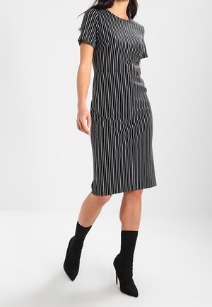 Donna che indossa un vestito nero al ginocchio con strisce verticali bianche e stivaletti calzino neri con tacco alto, che cammina in avanti.