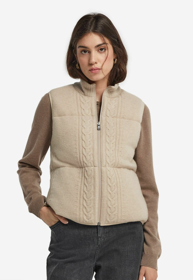 Beige stickad väst med hög krage, med ett kabelmönster fram, kompletterad med långa bruna ärmar. Dragkedja som stängning.