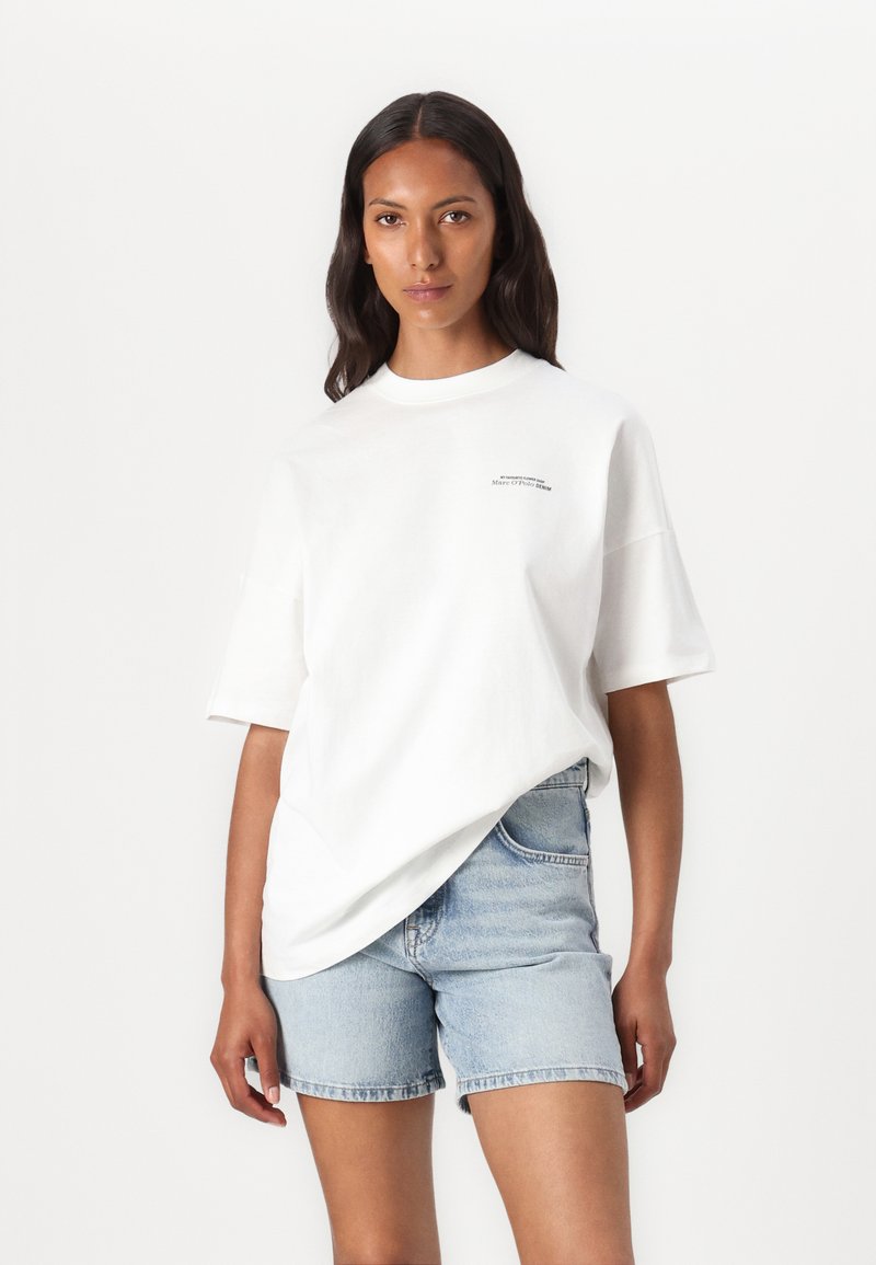 Weißes, übergroßes Baumwoll-T-Shirt mit kurzen Ärmeln und einem kleinen schwarzen Logo auf der Brust, kombiniert mit hellblauen Denim-Shorts.