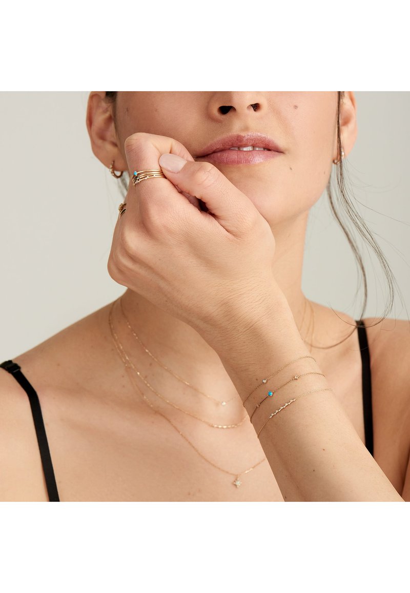 Das Goldschmuckset umfasst geschichtete Halsketten und Armbänder mit kleinen Akzenten, die einen blauen Stein und zarte geometrische Formen zeigen.