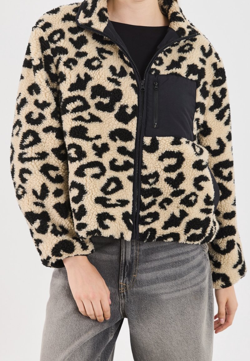 Beżowo-czarna polarowa kurtka w panterkę, z czarną kieszenią na suwak na klatce piersiowej oraz luźnym krojem i elastycznymi mankietami.