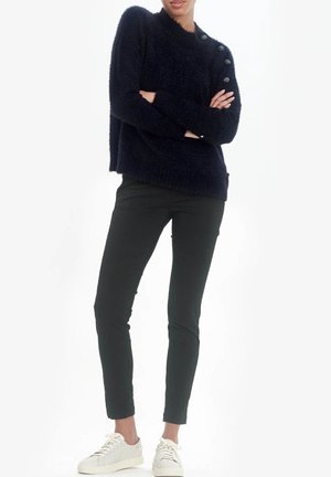 Personne debout, bras croisés, portant un pull noir en tissu duveteux avec des boutons et un pantalon noir taille fine, assortis à des baskets blanches.