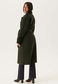 Cappotto di lana verde scuro con vita regolabile, colletto alto, maniche lunghe e taglio dritto, indossato sopra jeans scuri e stivali con tacco.