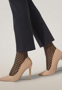 Nude högklackade skor med spetsiga tår, tillsammans med svarta fisknätsstrumpor. Byxorna är mörka med rak passform, vilket framhäver skorna.