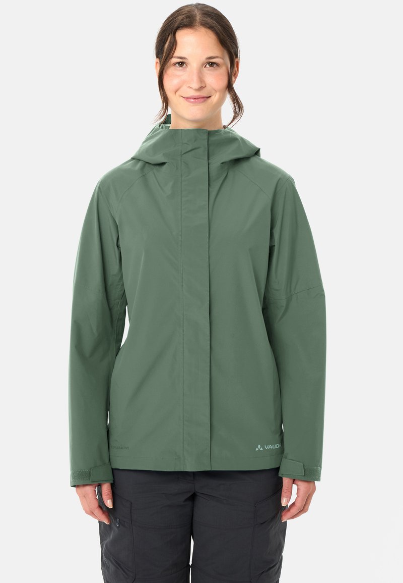 Frau trägt eine grüne, wasserabweisende Kapuzenjacke und schwarze Cargo-Hosen, steht vor einem einfarbigen hellen Hintergrund.