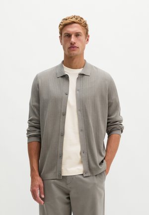 Mann mit lockigem blondem Haar, der eine hellgraue, gerippte, knöpfbare Strickjacke, ein weißes Hemd und graue Hose trägt, steht mit einer Hand in der Tasche.