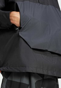 Giacca impermeabile nera con una texture liscia e un taglio distintivo, caratterizzata da una grande tasca laterale. Il design enfatizza la funzionalità.