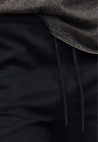 Μαύροι παντελόνες με ορατά κορδόνια σφιξίματος, σε συνδυασμό με γκρι ύφασμα από ένδυμα που βρίσκεται από πάνω.