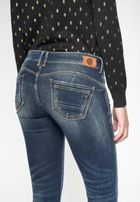 Jeans de mezclilla en azul oscuro con un aspecto desgastado. Presentan cinco bolsillos, costuras en contraste y una etiqueta de cuero marrón en la cinturilla.