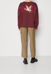 Bordeauxroter Sweatshirt mit großem beigem Grafikdruck, kombiniert mit khakifarbenen Cargo-Hosen mit Seitentaschen und schwarzen klobigen Schuhen.