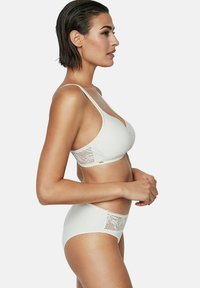 Femme aux cheveux noirs courts portant un soutien-gorge en dentelle blanc et une culotte haute assortie en dentelle, se tenant de profil devant un fond uni.