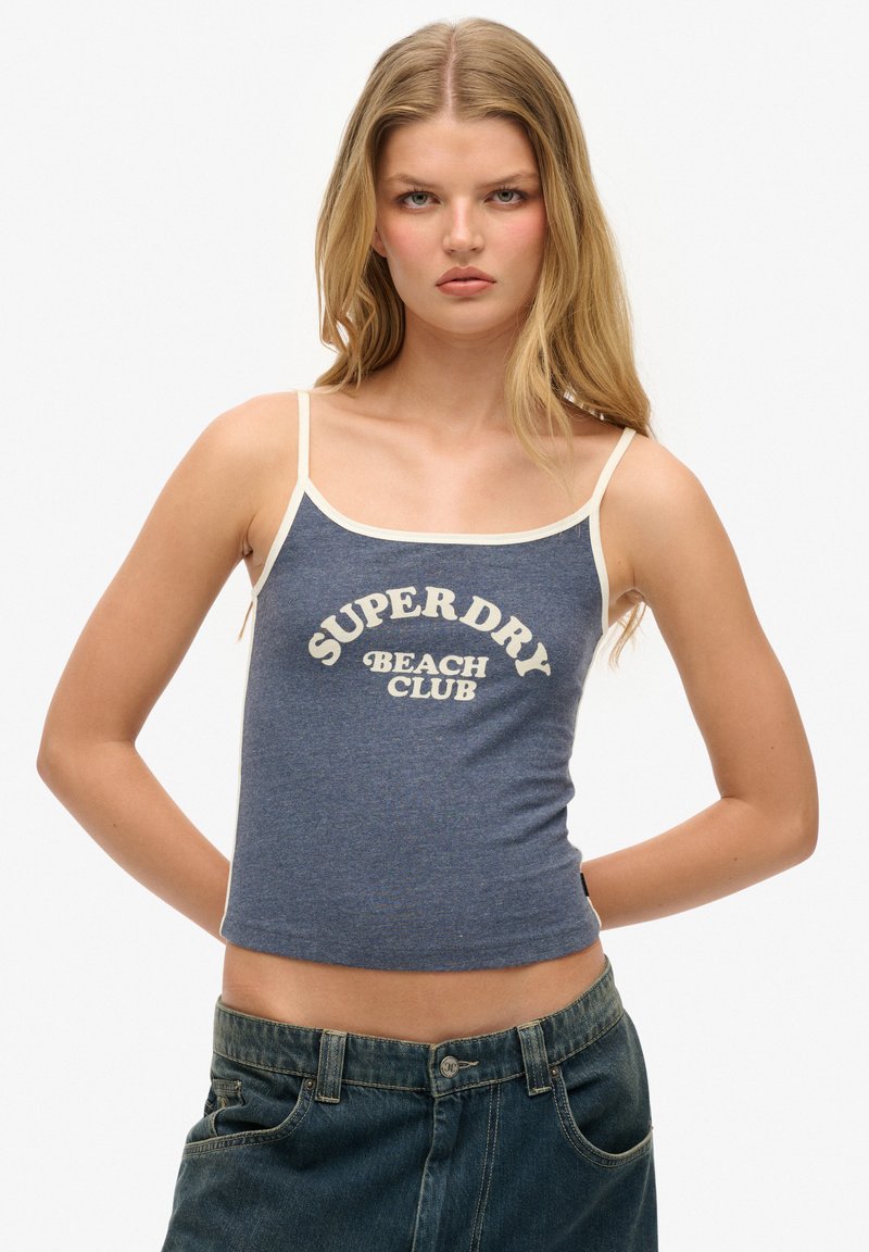Top de tirantes de algodón azul con correas color crema que presenta la impresión "SUPERDRY BEACH CLUB" en una fuente retro en el centro. Diseño ajustado, estilo casual.